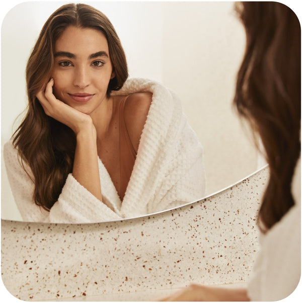 Woman in a white bathrobe looking at her reflection in a mirror while wondering what causes oily hair